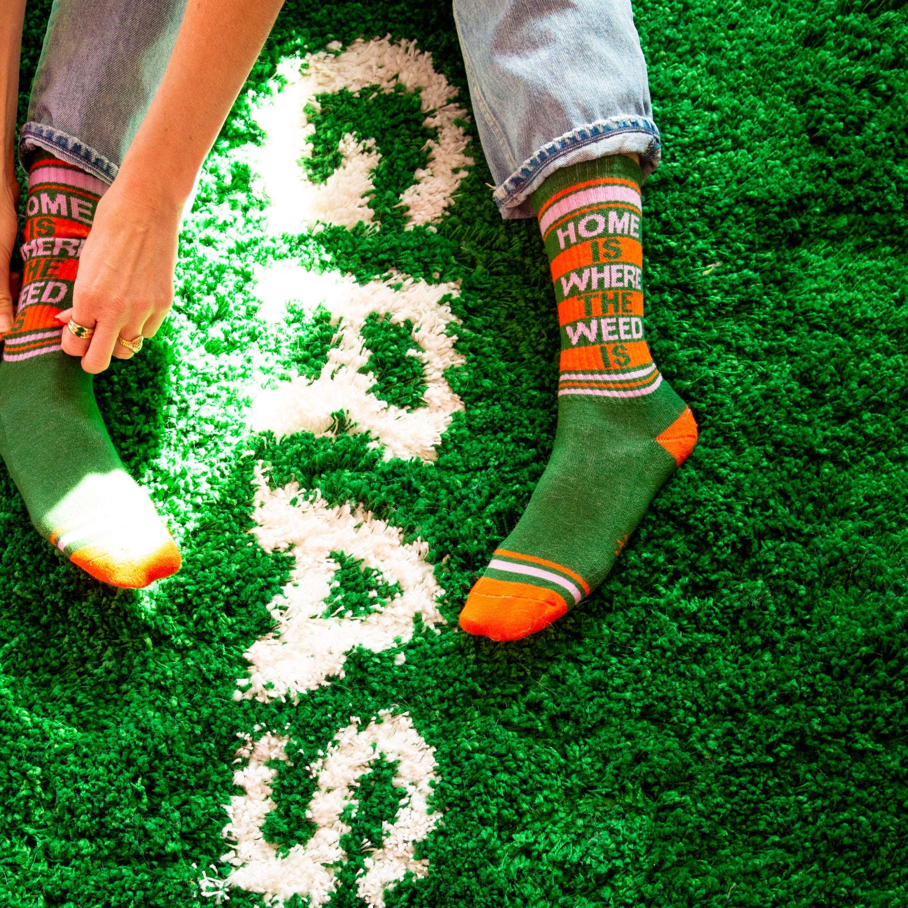 A pair of green, orange, and pink crew socks that read "home is where the weed is" with the Gumball Poodle logo on the arch.