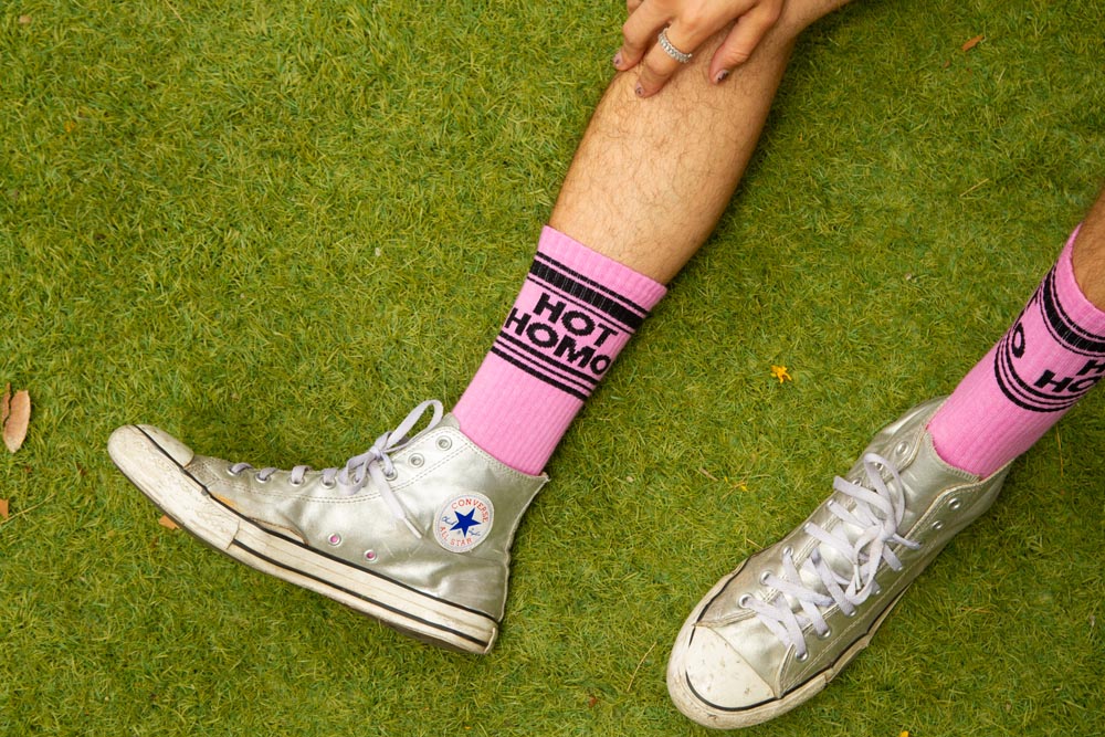 A pair of hot pink and black crew socks that read "hot homo" demonstrated with silver Converse.